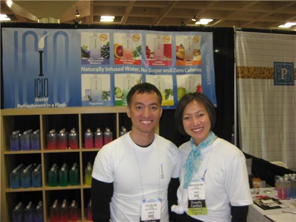 A smiling man and woman in white shirts stand together in front of an ICIO Water banner, with shelves of colorful flavored water bottles behind them.