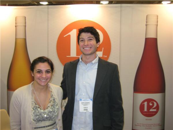 Justin Schwartz, identified by his badge, smiles beside a woman in a cream cardigan in front of a 12 booth with an orange logo and two tall bottles.