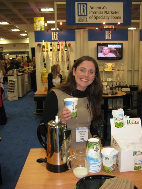 A smiling woman holds a PG2go paper cup at a table with a coffee percolator, a pitcher of milk and a box of PG2go cups, in front of the Liberty Richter booth.
