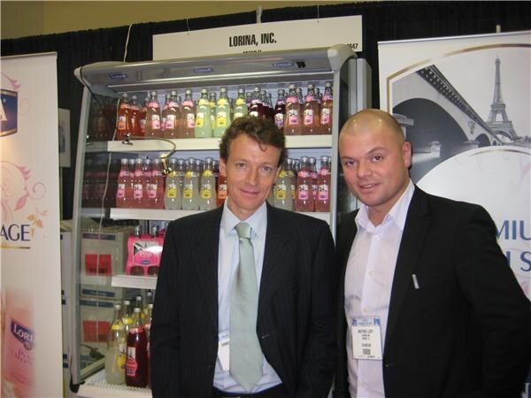 Two men in dark suits pose beside a refrigerated case of bottled lemonades and sodas under a Lorina, Inc. sign.