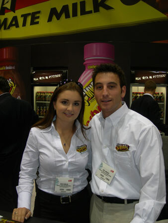 A woman and a man in white shirts with logo patches and name badges stand together in front of a large milk bottle display and refrigerated coolers under a sign reading 'Milk with an attitude'.