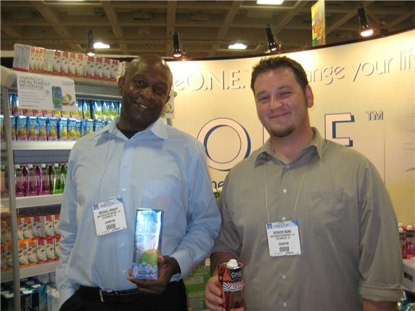 Michael Wright, left, holds a carton of O.N.E. coconut water beside a bearded man holding an O.N.E. drink; shelves of beverage cartons are behind them.