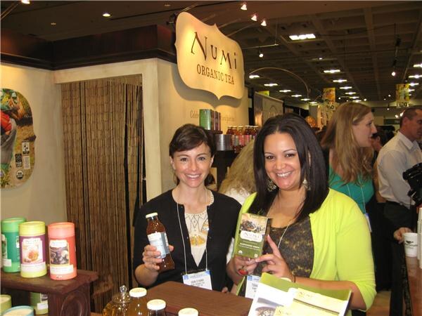 Two smiling women stand at a Numi Organic Tea booth; one holds a bottled tea and the other holds a box of tea, with tea tins on a shelf nearby.