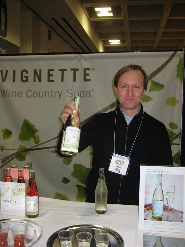Pat Galvin holds up a bottle at the Vignette Wine Country Soda booth, with bottles, a sign for California Brut and tasting cups on the table in front of him.