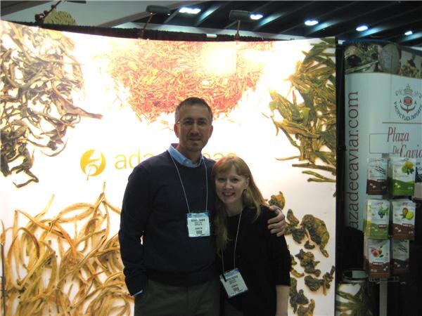 A man and a woman wearing badges pose together in front of a booth with tea leaf graphics, beside a Plaza de Caviar display of packaged products.