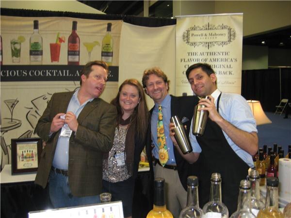 Three men and a woman pose at the Powell & Mahoney booth; one man holds two cocktail shakers, and bottles of mixers sit in the foreground.