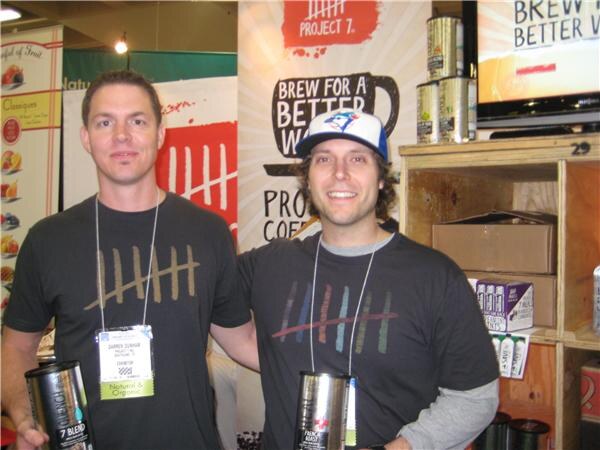 Two men in dark T-shirts with tally-mark designs stand in front of a Project 7 banner, each holding a can; one wears a blue Toronto Blue Jays cap.