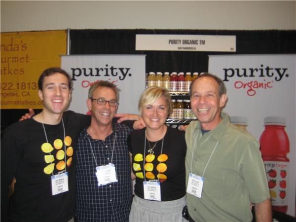 Four smiling people, two in black T-shirts with yellow fruit designs, stand arm in arm between Purity Organic banners, with bottled juices on shelves behind them.