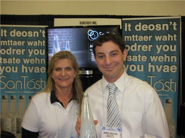 A woman and a man in white shirts stand at the SanTásti booth, the man holding a clear glass bottle, with SanTásti banners and a screen behind them.