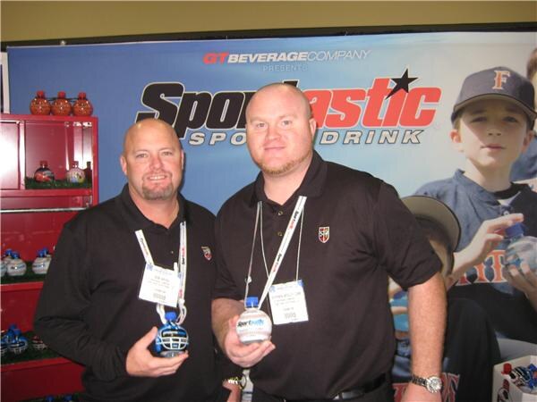 Two men in black polo shirts and badges hold small helmet-shaped Sportastic bottles in front of a GT Beverage Company Sportastic Sports Drink banner showing a boy in a cap.