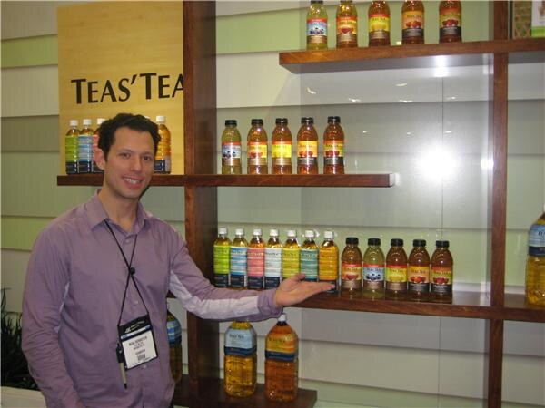 A smiling man in a purple shirt gestures toward wooden shelves of bottled tea beneath a Teas' Tea sign.
