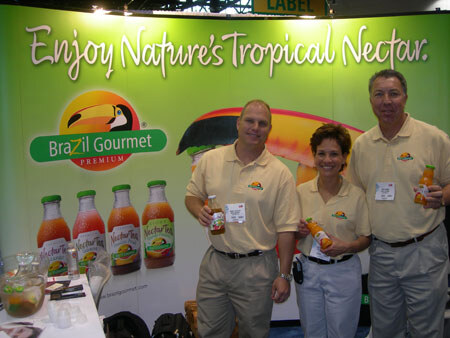 Two men and a woman in matching tan shirts hold bottles of Nectar Tropical in front of a Brazil Gourmet backdrop reading 'Enjoy Nature's Tropical Nectar' with bottles pictured on it.