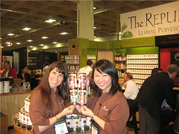 Two smiling women in brown uniforms hold tins of tea at The Republic of Tea booth, with shelves of tea tins and a company banner behind them.