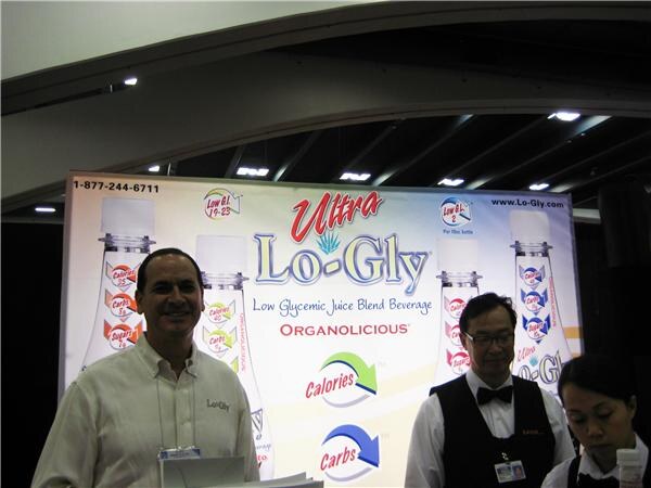 A smiling man in a cream shirt stands in front of an Ultra Lo-Gly backdrop; a man in a vest and bow tie and a woman stand to his right.