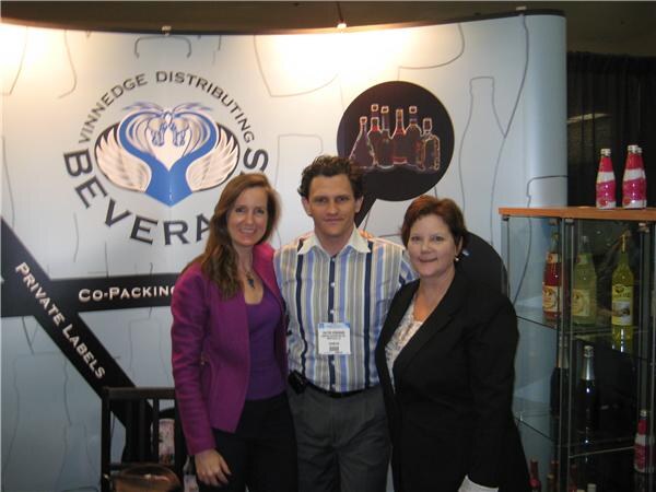 Two women and a man stand together in front of a Vinnedge Distributing banner mentioning private labels and co-packing, beside a display case of bottles.