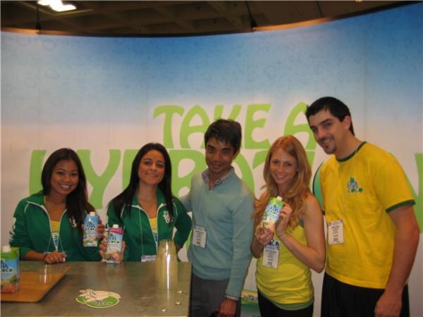 Five people in green and yellow Vita Coco clothing stand behind a counter, two women holding cartons of coconut water, in front of a blue and green backdrop.