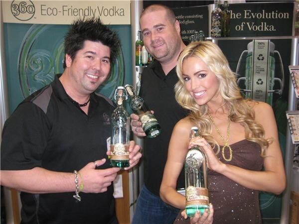 Two men and a blonde woman hold bottles of 360 Vodka in front of banners reading Eco-Friendly Vodka and The Evolution of Vodka.
