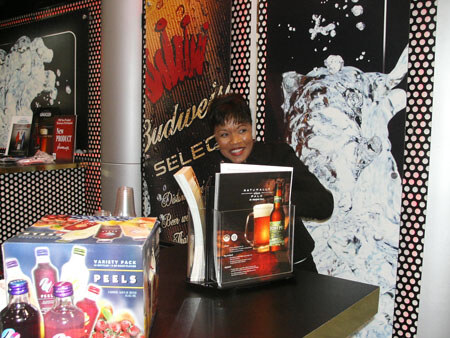 A smiling woman in a dark top stands behind a counter in front of Budweiser Select signage, with a Peels variety pack box and a brochure holder with a beer glass and bottle on the counter.