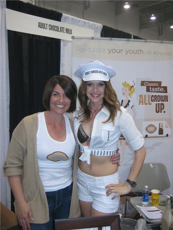 A woman in a tank top and cardigan stands beside a woman in a white outfit and Adult Chocolate Milk cap, in front of an Adult Chocolate Milk banner.