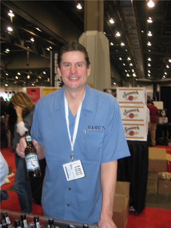A smiling man in a blue Bard's shirt and lanyard holds a bottle of Bard's beer, with stacked Sonwerk boxes and a convention hall behind him.