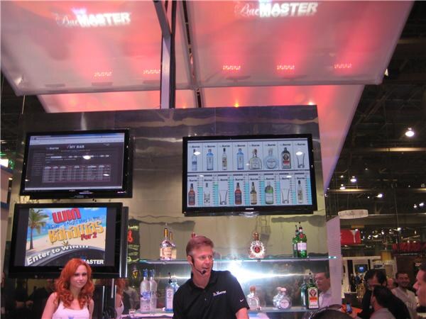 A man in a black shirt with a headset stands at a Bar Master display with liquor bottles on shelves and screens above. A red-haired woman stands at left, and a screen shows a contest promotion.