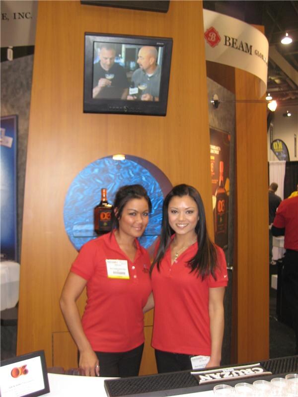 Two women in red polo shirts stand at the Beam Global booth, with a Cruzan bar mat in front and a wall-mounted screen and bottle display behind them.