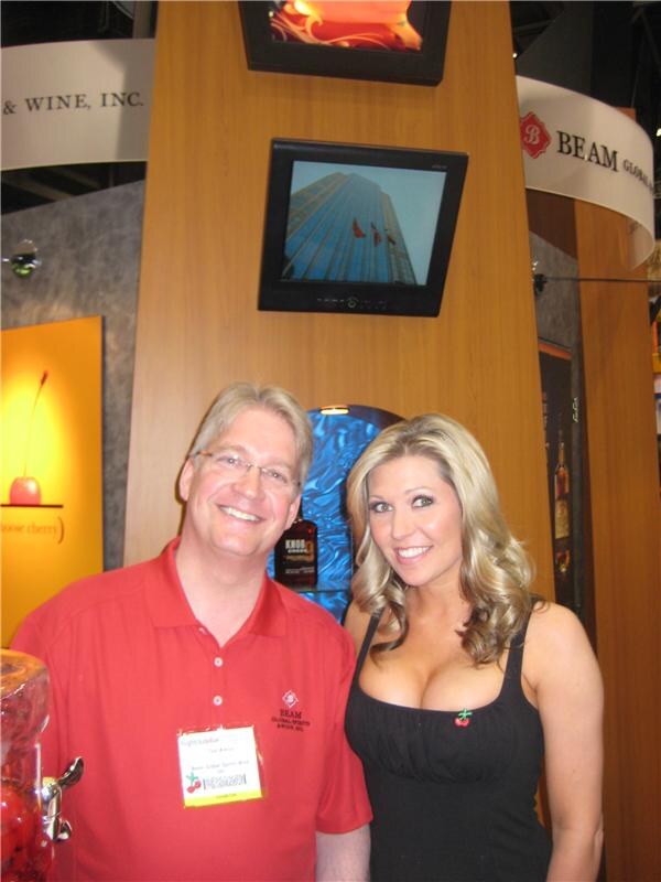 A smiling man with glasses in a red Beam polo and a smiling woman with long wavy blonde hair in a black top pose in front of the Beam Global Spirits & Wine booth with display screens.