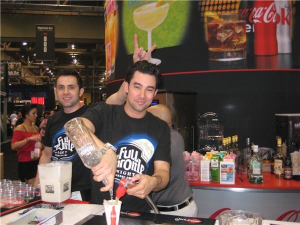 Two smiling men in black Full Throttle shirts stand behind a bar. One pours a drink from a bottle into a Coca-Cola cup. Coca-Cola signage hangs behind them.