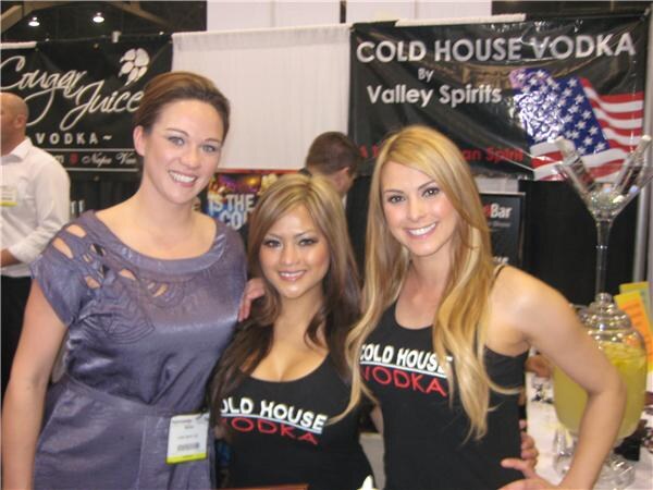 Three smiling women pose in front of a Cold House Vodka by Valley Spirits banner. Two wear black Cold House Vodka tops. A Cougar Juice Vodka sign appears at left.