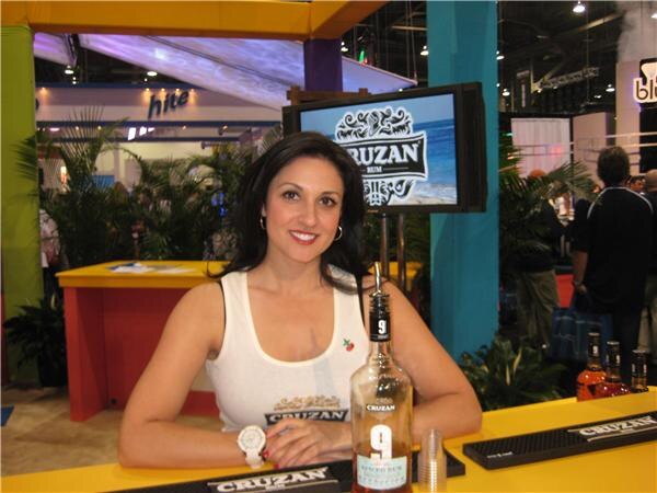 A smiling woman with dark hair in a white Cruzan tank top stands at a yellow bar with a bottle of Cruzan rum. A screen behind her shows the Cruzan logo.