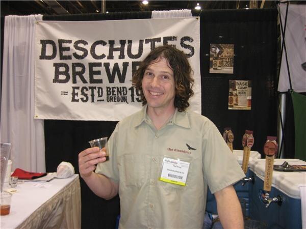 A smiling man with curly dark hair in a tan work shirt holds a small cup of beer in front of a Deschutes Brewery banner. Deschutes tap handles are on the table behind him.