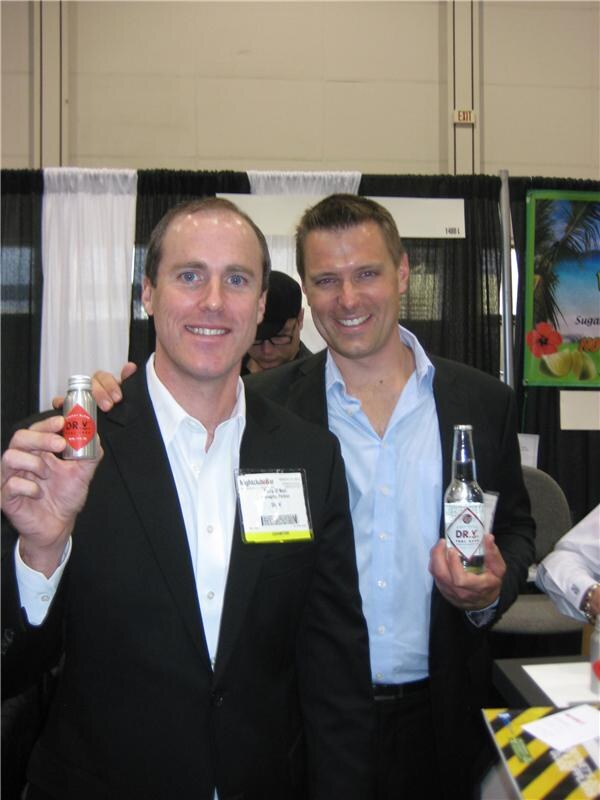 Two smiling men in dark jackets pose at a booth. The man at left holds a small Dr. V aluminum bottle and the man at right holds a Dr. V glass bottle.