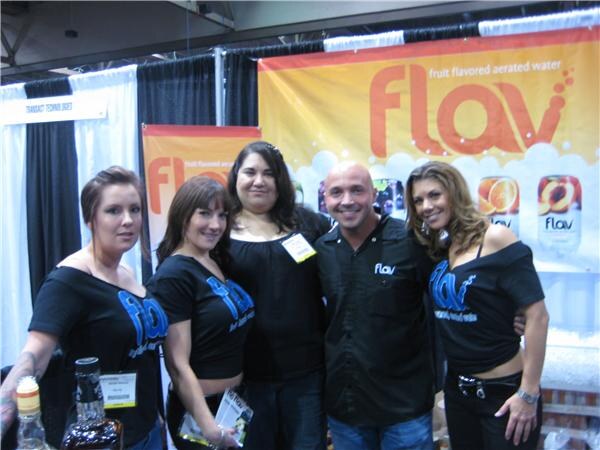 Five smiling people, four of them in black Flav shirts, pose in front of an orange Flav fruit flavored aerated water banner at a trade-show booth.