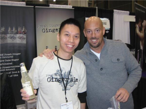 A smiling man in a Fresh Ginger Ale T-shirt holds a bottle of ginger ale while a bald man with a goatee in a gray sweater stands beside him, in front of ginger ale banners.
