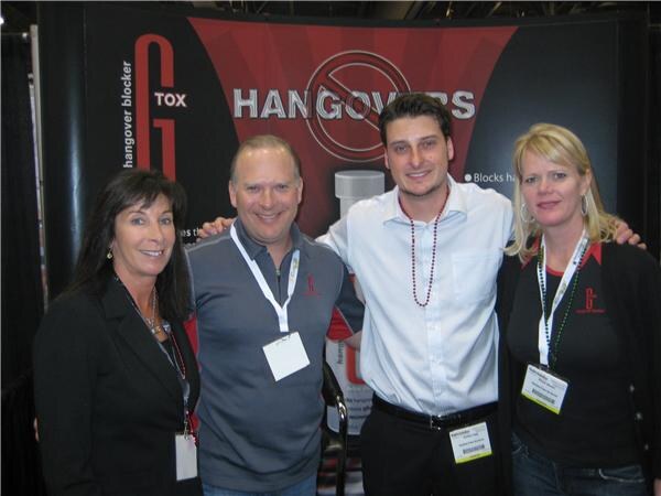 Two women and two men pose together in front of a G Tox hangover blocker booth display with the words Hangovers. All four wear trade-show badges on lanyards.
