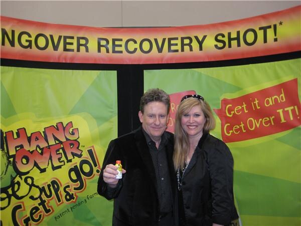 A man holding a small bottle and a woman with sunglasses on her head stand in front of a green and yellow Hangover Recovery Shot banner reading Get it and Get Over IT.