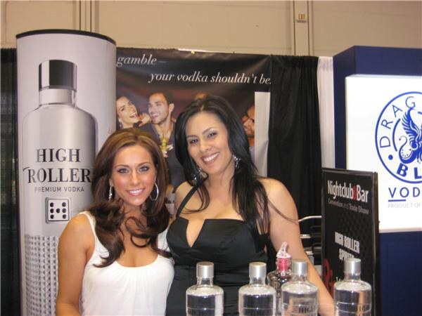 Two smiling women stand in front of a High Roller Premium Vodka display, with clear bottles in front of them and a Dragon Blue Vodka sign at right.