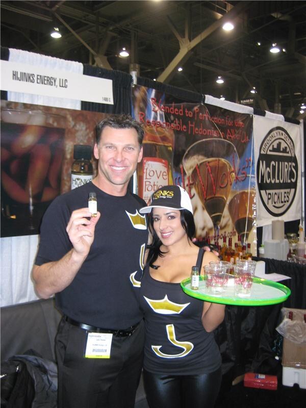 A man holding a small bottle and a woman in a Hijinks cap holding a tray of cups stand in the Hijinks Energy booth, with Love Potion and McClure's Pickles banners behind them.