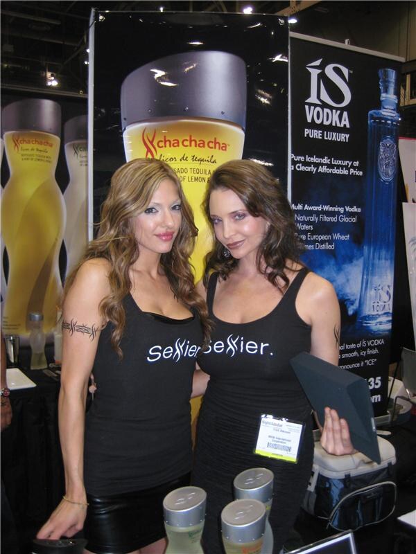 Two women in black Sexier tank tops pose in front of a Cha Cha Cha tequila banner and an IS Vodka banner showing a blue bottle.