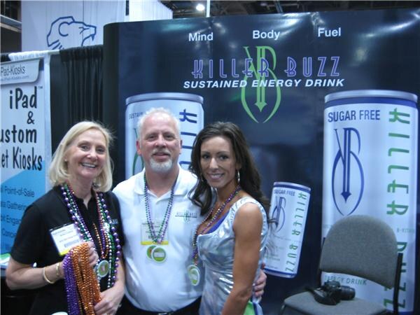 Two women and a bearded man wearing Mardi Gras beads stand in front of a Killer Buzz sustained energy drink display showing sugar-free cans.