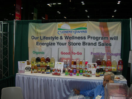 A Clement Pappas booth with a banner reading 'Our Lifestyle & Wellness Program will Energize Your Store Brand Sales' and a table of juice bottles, with a seated man at the table's edge.