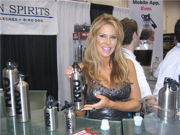 A smiling woman with long blonde hair holds a metal KRU bottle at a glass counter displaying several more KRU bottles, with people behind her.