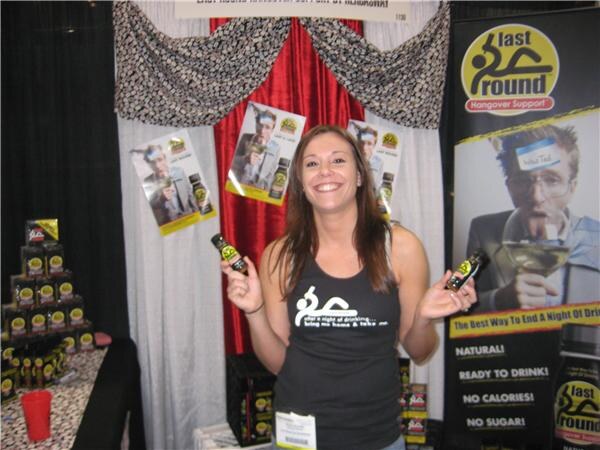A smiling woman holds two small bottles in the Last Round Hangover Support booth, with product stacks, posters and a tall Last Round banner behind her.