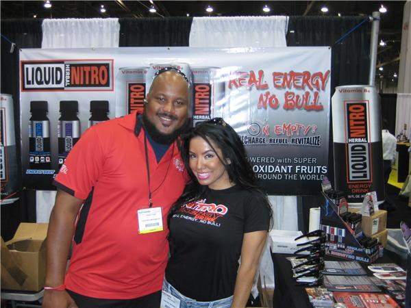 A man in a red shirt and a woman in a black Nitro shirt pose in front of a Liquid Herbal Nitro banner reading Real Energy No Bull.