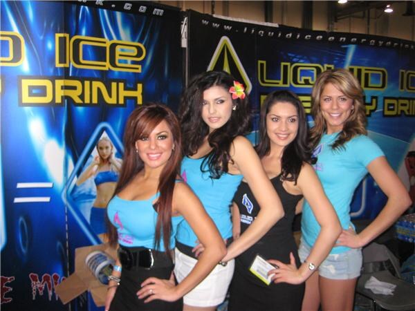 Four women in light blue and black outfits pose close together in front of Liquid Ice Energy Drink banners with blue and yellow lettering. One woman wears a pink flower in her hair.