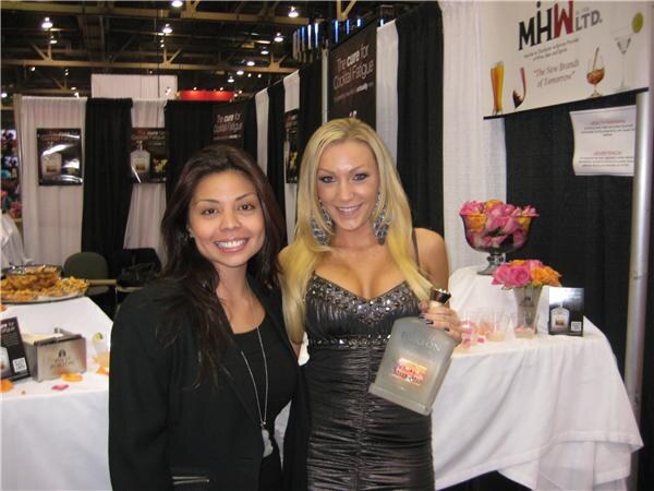 Two smiling women stand at a draped booth with the MHW Ltd. sign overhead; one holds a bottle, and pink and orange flowers sit on a white-clothed table beside them.