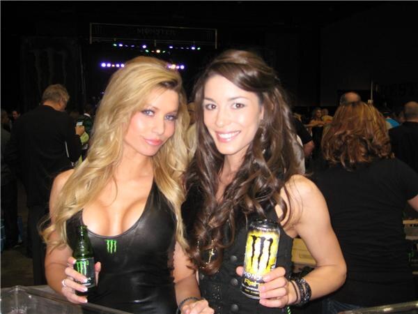 Two women pose close together, one with long blonde hair holding a green Monster Energy bottle and the other with long brown hair holding a yellow Monster Rehab can. Attendees are behind them.