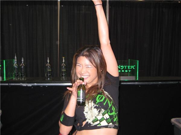 A smiling woman with one arm raised holds a small Monster Energy bottle. Behind her is a bar with liquor bottles and a green Monster sign against black curtains.