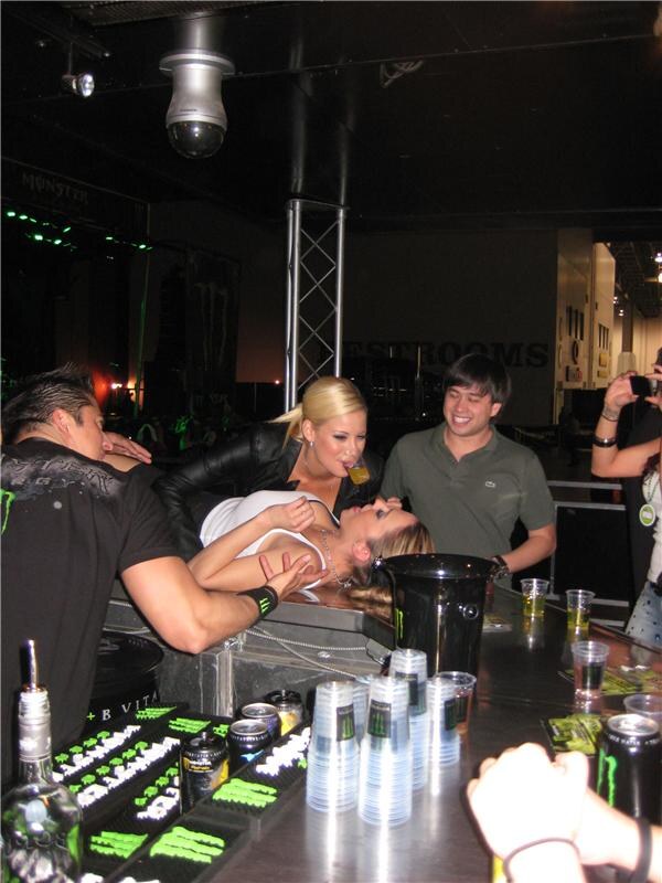 At a Monster Energy bar, a woman leans back across the counter while a blonde woman pours a drink toward her mouth. A smiling man in a green polo with a Lacoste logo watches, with cups and cans on the bar.