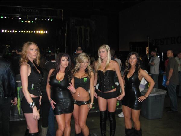 Five women in black outfits, some with Monster Energy logos, stand in a row in a dim hall. A stage with lights is behind them and attendees stand nearby.